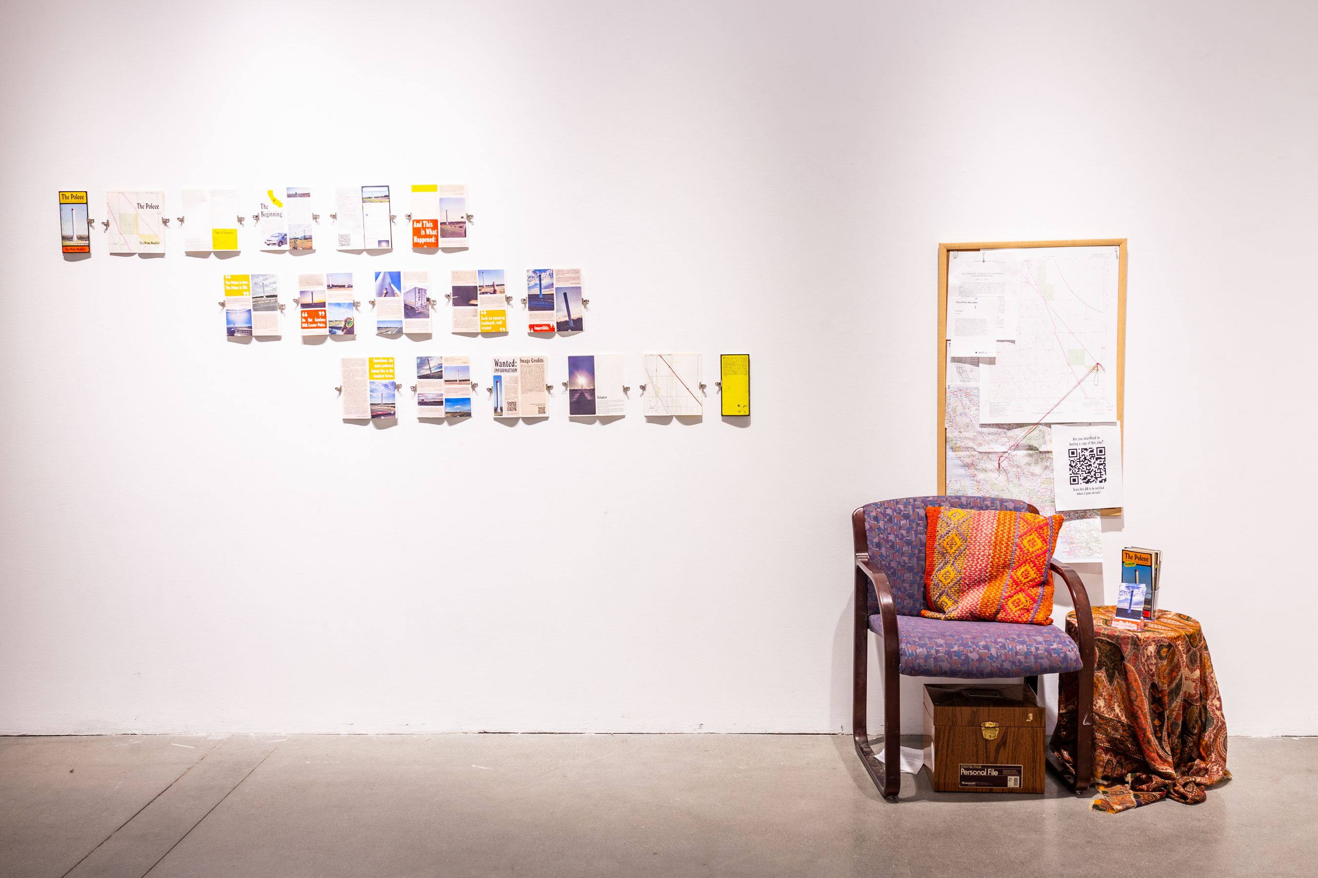 A chair sits in the center of the frame, with a pillow on it. On the right of the chair is a small circular coffee table with brochures. In the back is a white wall with a corkboard with maps and diagrams.